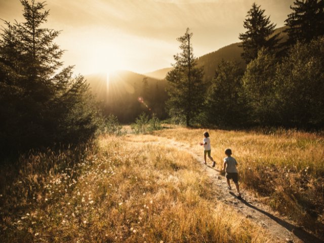 duas crianças fotografadas correndo em um campo com gramas marrons ao por do sol. 