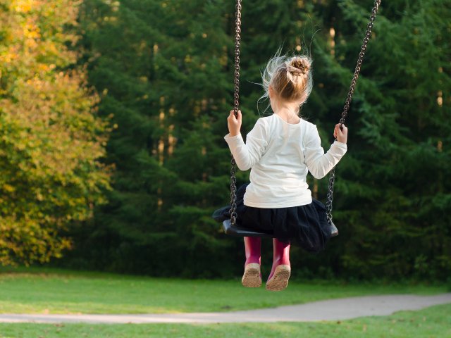 Criança fotografada de costas enquanto se balança em um balanço no parque
