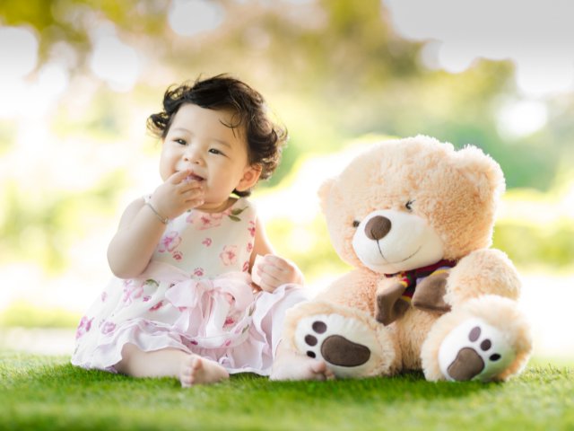 Criança ao lado de um ursinho de pelúcia sentada na grama verde
